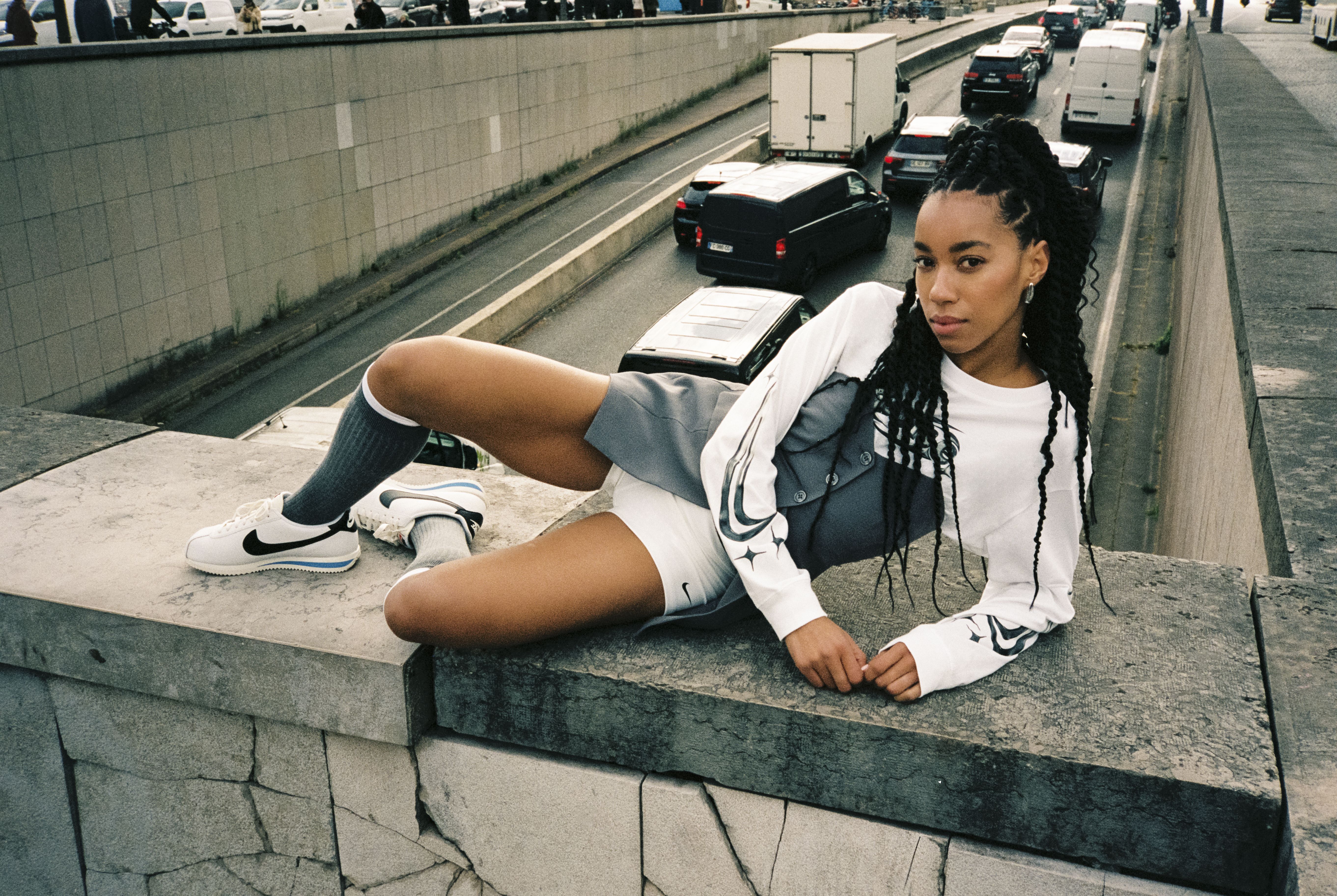 Sieviete baltā NIKE sporta kreklā un baltos NIKE apavos pozē uz betona sienas virs ielas